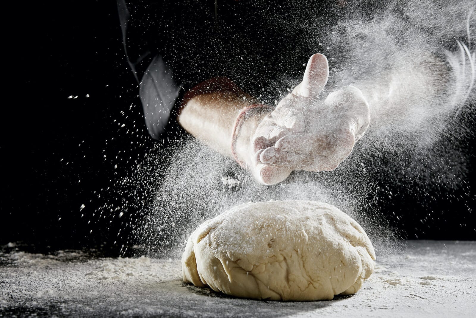 Unidentified man wearing black chef suit hidden in the shadows sprinkling white flour over brightly lit blob of dough